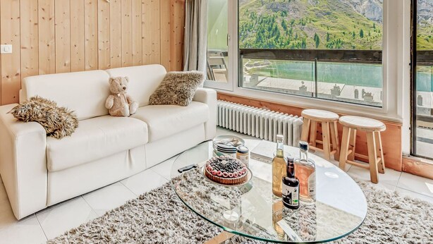 A modern living room with a white sofa, a glass coffee table, and a view of a lake with mountains in the background.