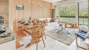 A modern kitchen with wooden accents, a dining area with a wooden table and chairs, a cozy seating area with a white sofa, and large windows offering a scenic view.