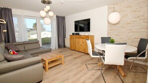 Un soggiorno moderno con divano, tavolino e zona pranzo con sedie. C'è una TV montata a parete e un mobile contenitore in legno.
