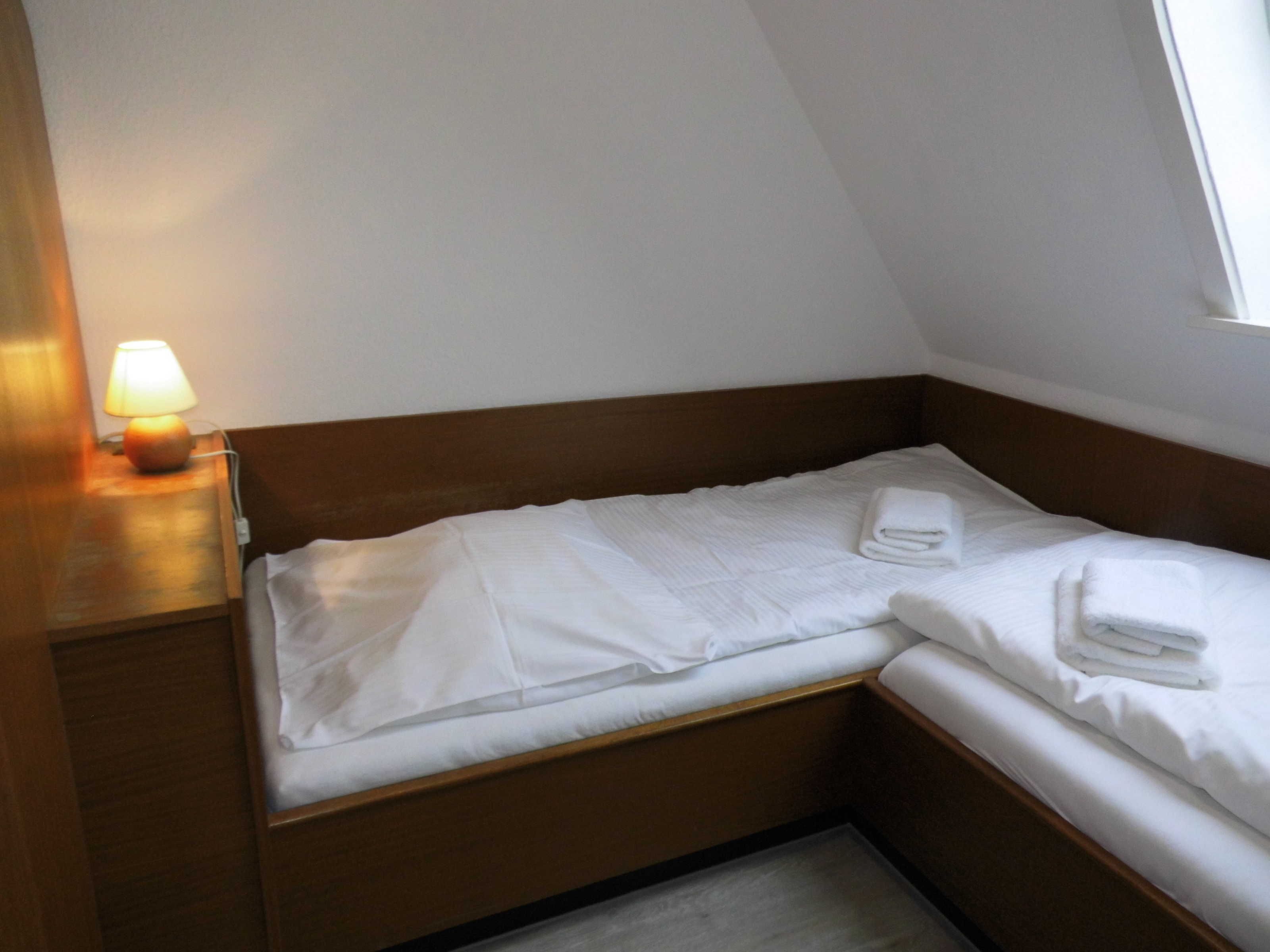 Habitación pequeña con una cama individual, cabecero de madera, lámpara de noche y ventana que deja entrar luz natural.