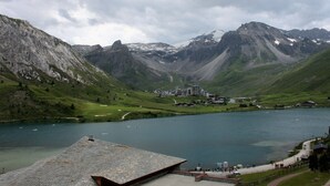 Paisaje montañoso con un lago, edificios y una zona pavimentada con gente.