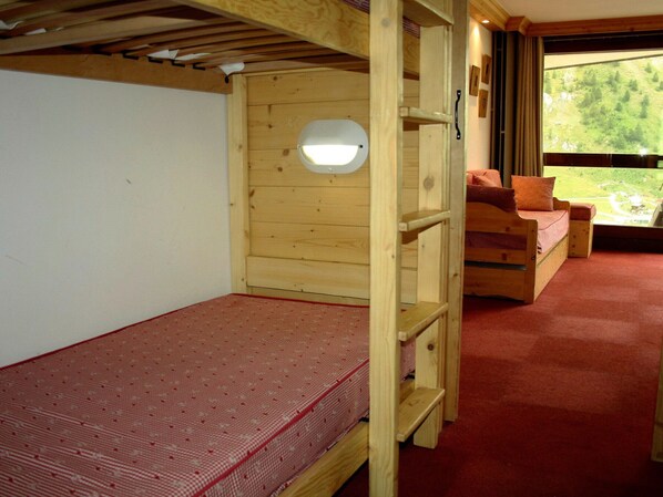 Ein hölzernes Etagenbett mit roter Matratze, ein kleines Fenster und Blick auf eine grüne Landschaft durch eine Glastür.