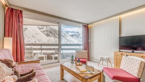 Una sala de estar acogedora con un sofá, una mesita de centro con aperitivos, un televisor y vistas a montañas nevadas.