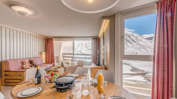 Un comedor con una mesa de madera lista para una comida, un sofá con cojines y vistas a un paisaje montañoso nevado a través de amplios ventanales.