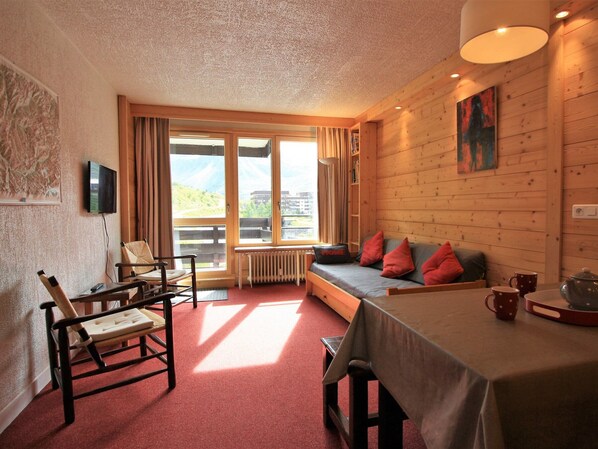 A cozy room with a wooden interior, a sofa with red cushions, a dining table, and a television mounted on the wall. - Renovated Apartment in Tignes - 6 Guests (Tignes-Le Lavachet)