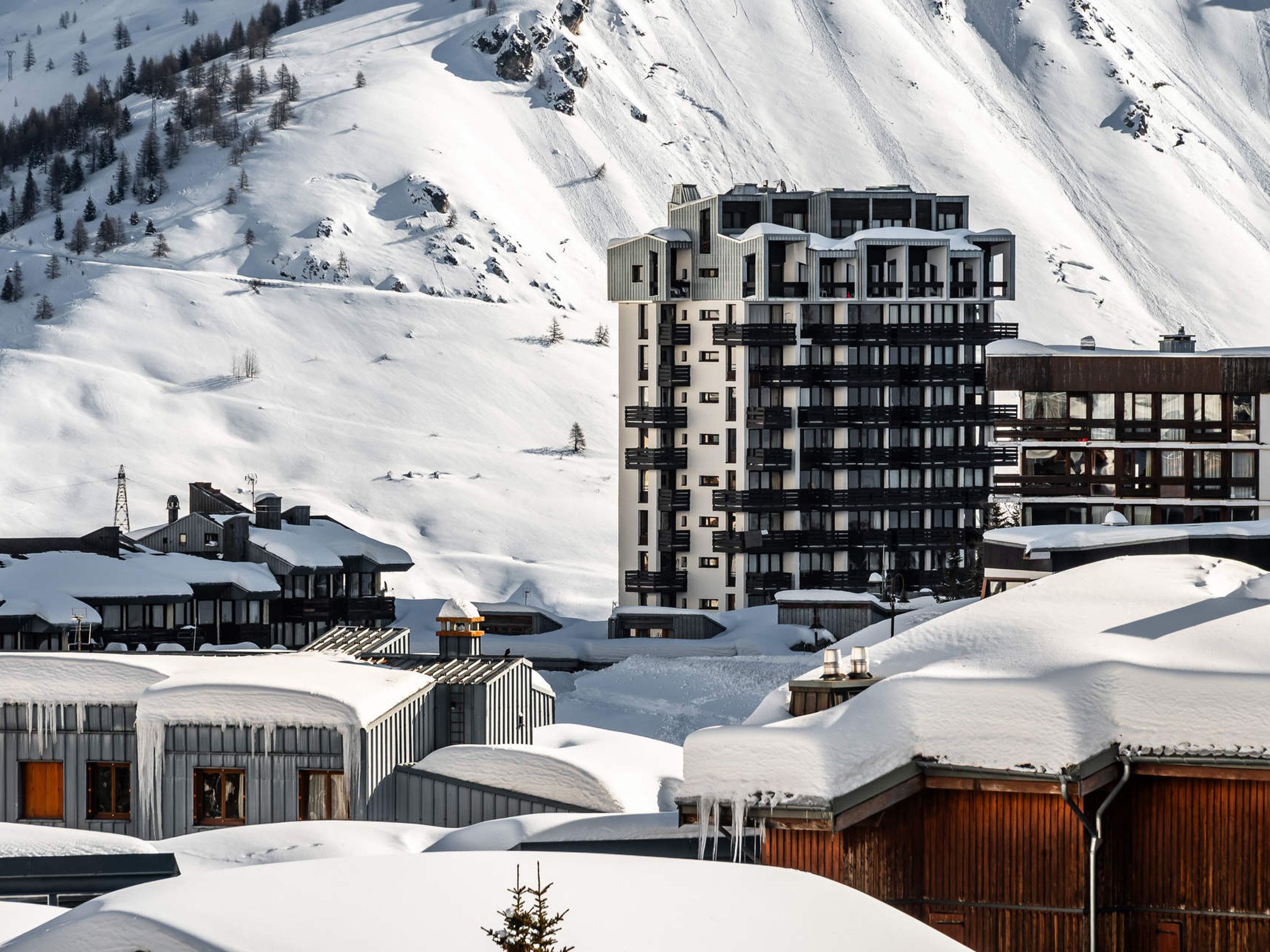 A snow-covered mountain town with modern buildings, including a multi-story hotel with balconies and a ski resort in the background.