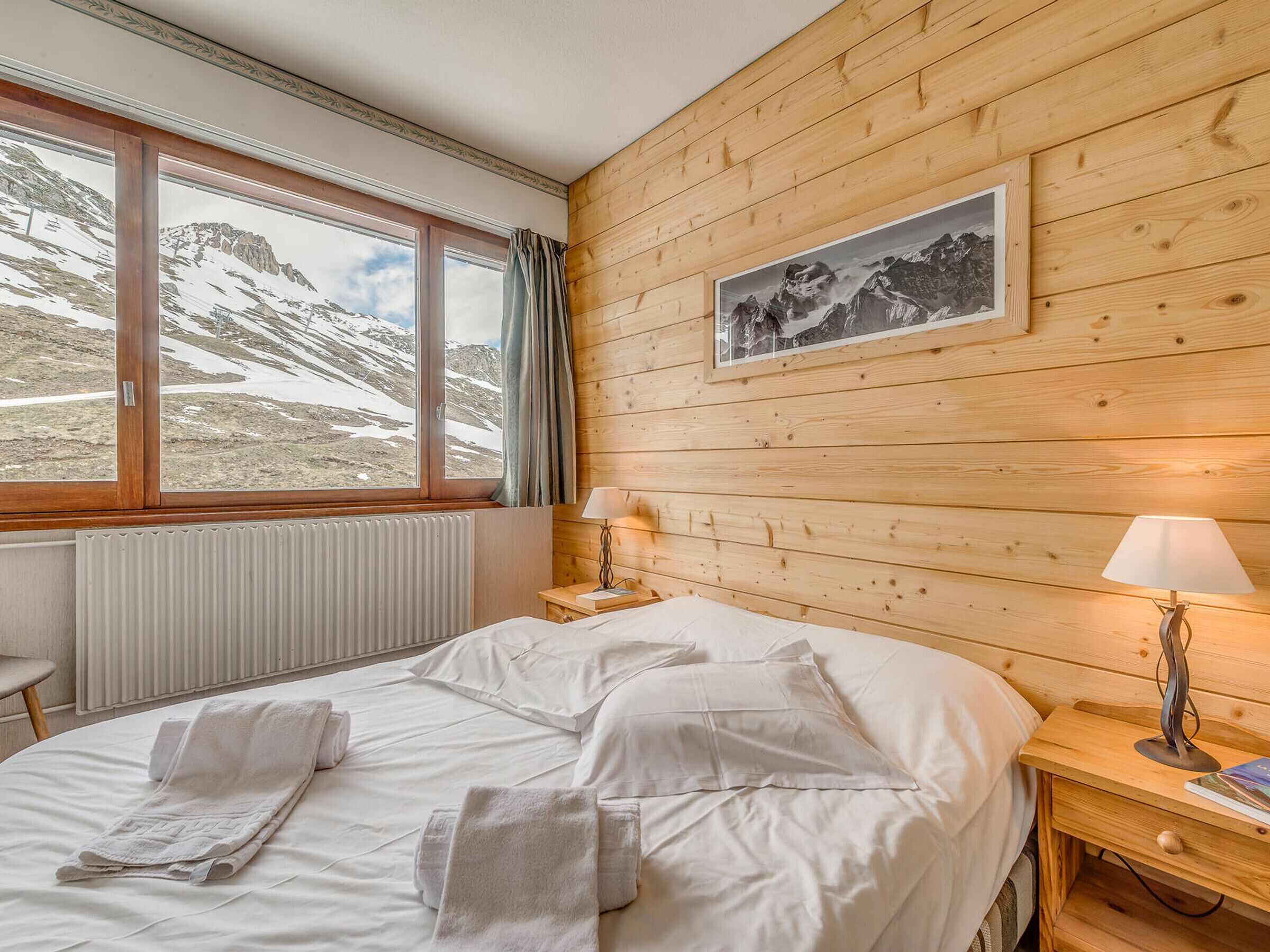 A hotel room with a wooden interior, a bed with white linens, a window with a mountain view, and a framed picture on the wall.