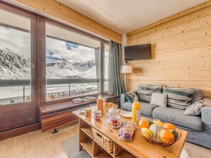 A cozy living room with a wooden interior, a sofa, a coffee table with breakfast items, and a view of a snowy mountain landscape through large windows. - Renovated 3* Apartment with WiFi, ski-in/ski-out, 6 people (Tignes)