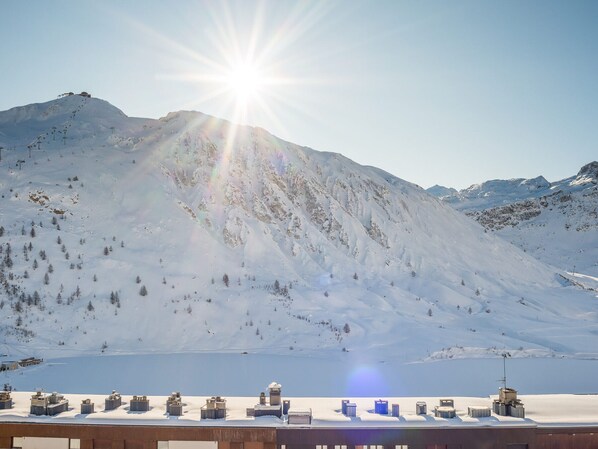 A snowy mountain landscape with a clear sky, a lake, and a row of buildings with a flat roof.