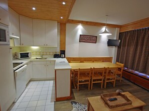 Una cocina funcional con armarios blancos, microondas, tostadora y cafetera. Cuenta con una mesa de comedor de madera con sillas, televisor montado en la pared y una cortina estampada.