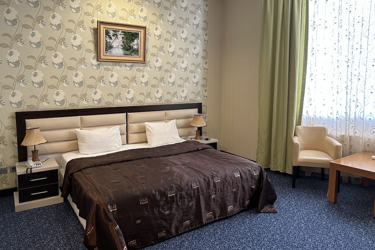 Ein Hotelzimmer mit einem großen Bett, zwei Nachttischen, einem Sessel und einem Fenster mit Vorhängen.
