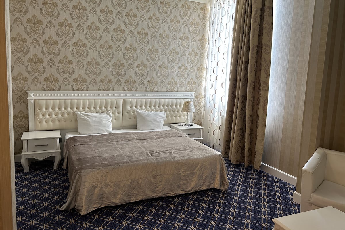 Ein Hotelzimmer mit einer gepolsterten Kopfteil, einem großen Bett, einem Nachttisch mit Lampe und einem gemusterten Teppich.