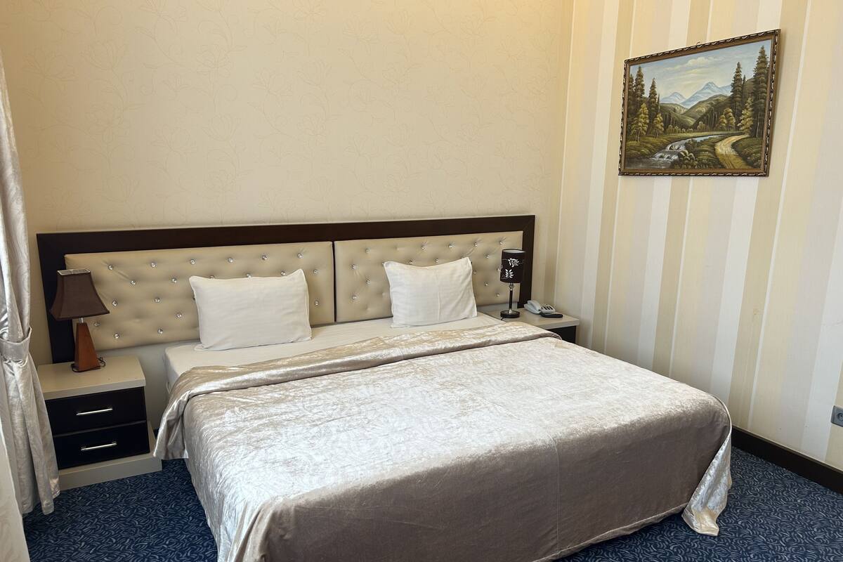 Ein Hotelzimmer mit einem großen Bett, einem Nachttisch, einer Lampe, einem Bild an der Wand und einem blau gemusterten Teppich.