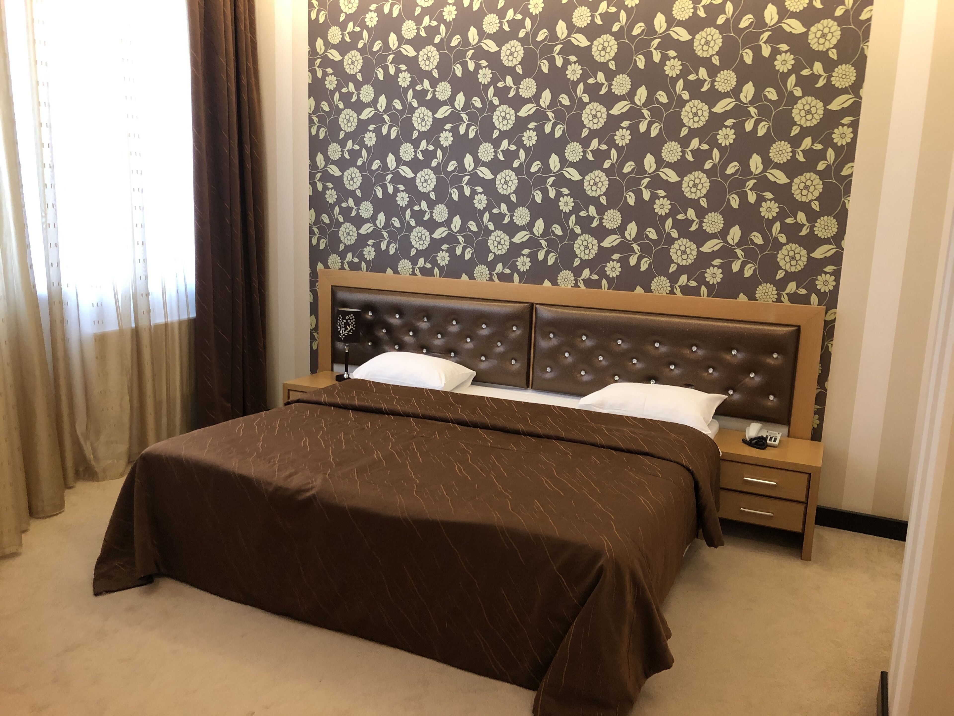 Habitación de hotel con una cama grande, un cabecero de madera, una mesita de noche y un papel tapiz floral en la pared.