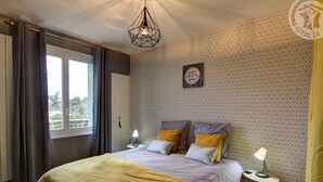 A bedroom with a bed, two bedside lamps, a geometric-patterned wallpaper, a window with curtains, and a ceiling light fixture.