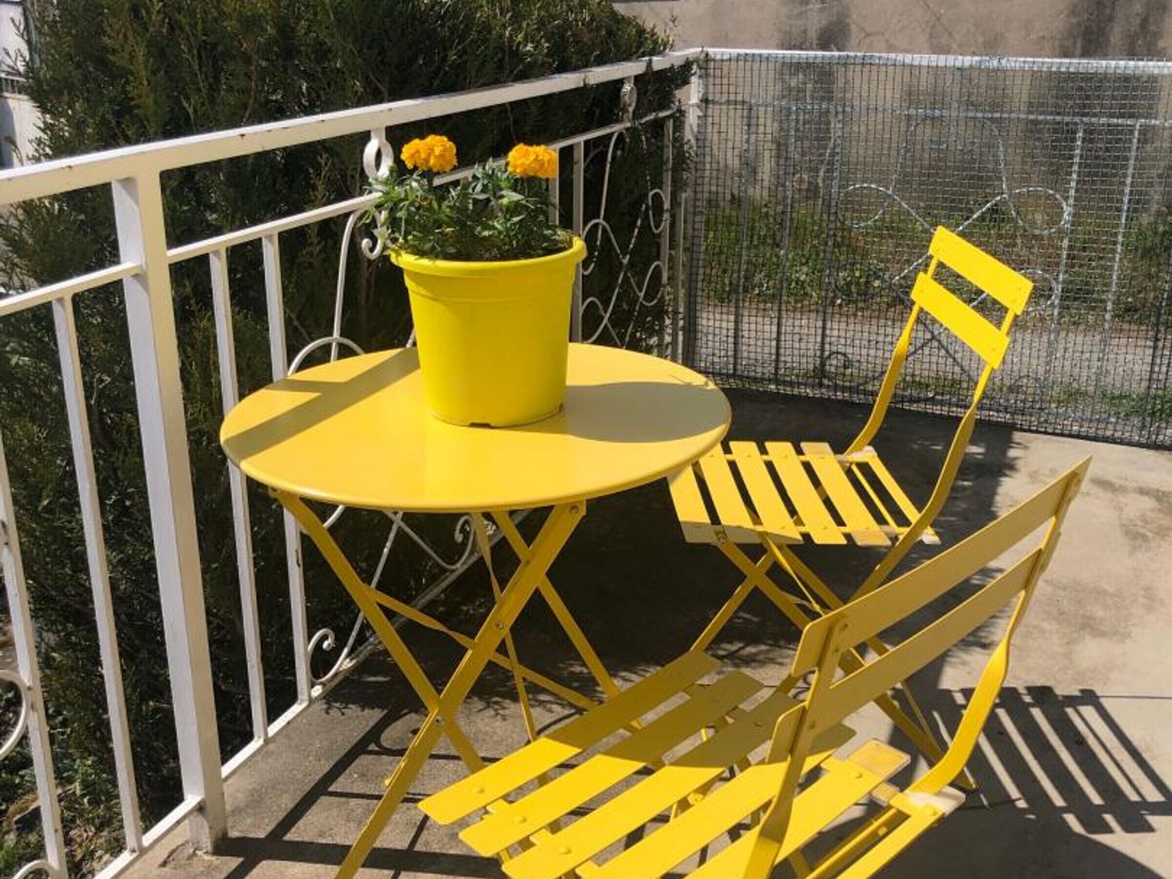 A yellow table and chair set on a balcony with a white railing and a potted plant with yellow flowers.