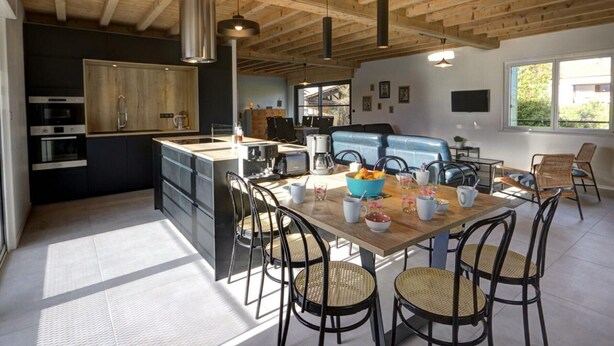 A modern kitchen with a central island, wooden ceiling beams, and a dining area with wicker chairs and a wooden table.