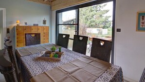 A dining area with a table set for two, a wicker chair, a wooden cabinet, and a window with a view of the outdoors.