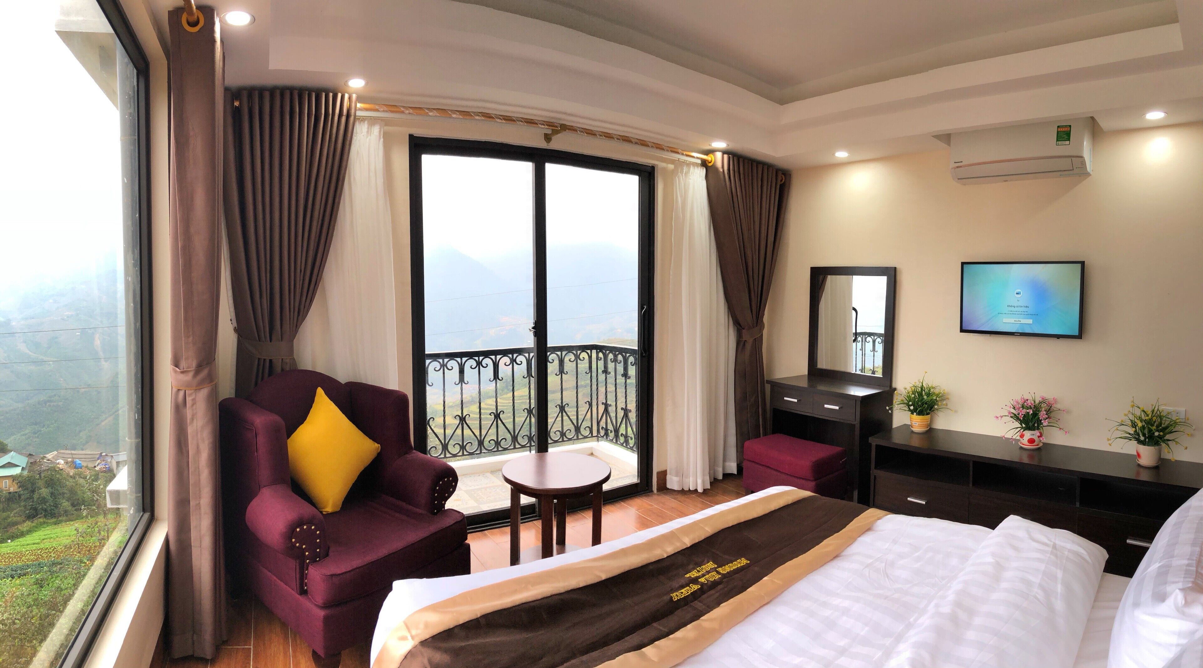 Habitación de hotel con cama, sofá, una mesita, escritorio, televisor y balcón con vistas a las montañas.