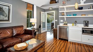 A modern living room with a leather sofa, a glass coffee table, and a dining area with white cabinetry and a kitchen island. - Pecan Villa with Access to Swimming Pool and Hot Tub (Fredericksburg)