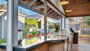 A covered outdoor patio with a grill, seating area, and a view of a house. - Pecan Villa with Access to Swimming Pool and Hot Tub (Fredericksburg)
