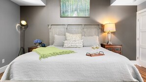 A bedroom with a large bed, a nightstand with a lamp, a wall-mounted painting, and a bedside table with a vase of flowers. - Pecan Villa with Access to Swimming Pool and Hot Tub (Fredericksburg)