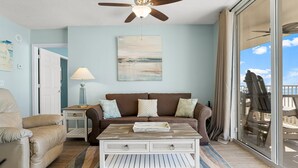 A living room with a ceiling fan, a brown sofa, a beige armchair, a coffee table, and a view of the outdoors through a sliding glass door. - Gulf-View King Bedroom | Beach Chairs Included | Long Beach Tower 3 (Panama City Beach)