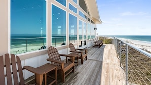 En træterrasse med Adirondack-stole, store vinduer med udsigt over stranden og et gelænder langs kanten.