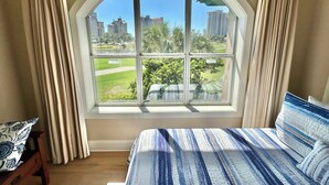 Une chambre dotée d’une grande fenêtre en arc de cercle, d’un lit avec une literie à rayures et offrant une vue sur un parc et des immeubles.