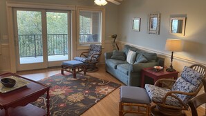 A living room with a sofa, chairs, a small table, and a floor lamp. There are large windows with blinds, a ceiling fan, and a decorative mirror on the wall.