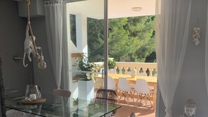 A modern dining area with a glass table, white chairs, and a view of a balcony with outdoor furniture.