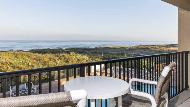 Un balcón con una mesa blanca y sillas de mimbre, con vistas a un paisaje costero y una vista despejada del mar y colinas lejanas.