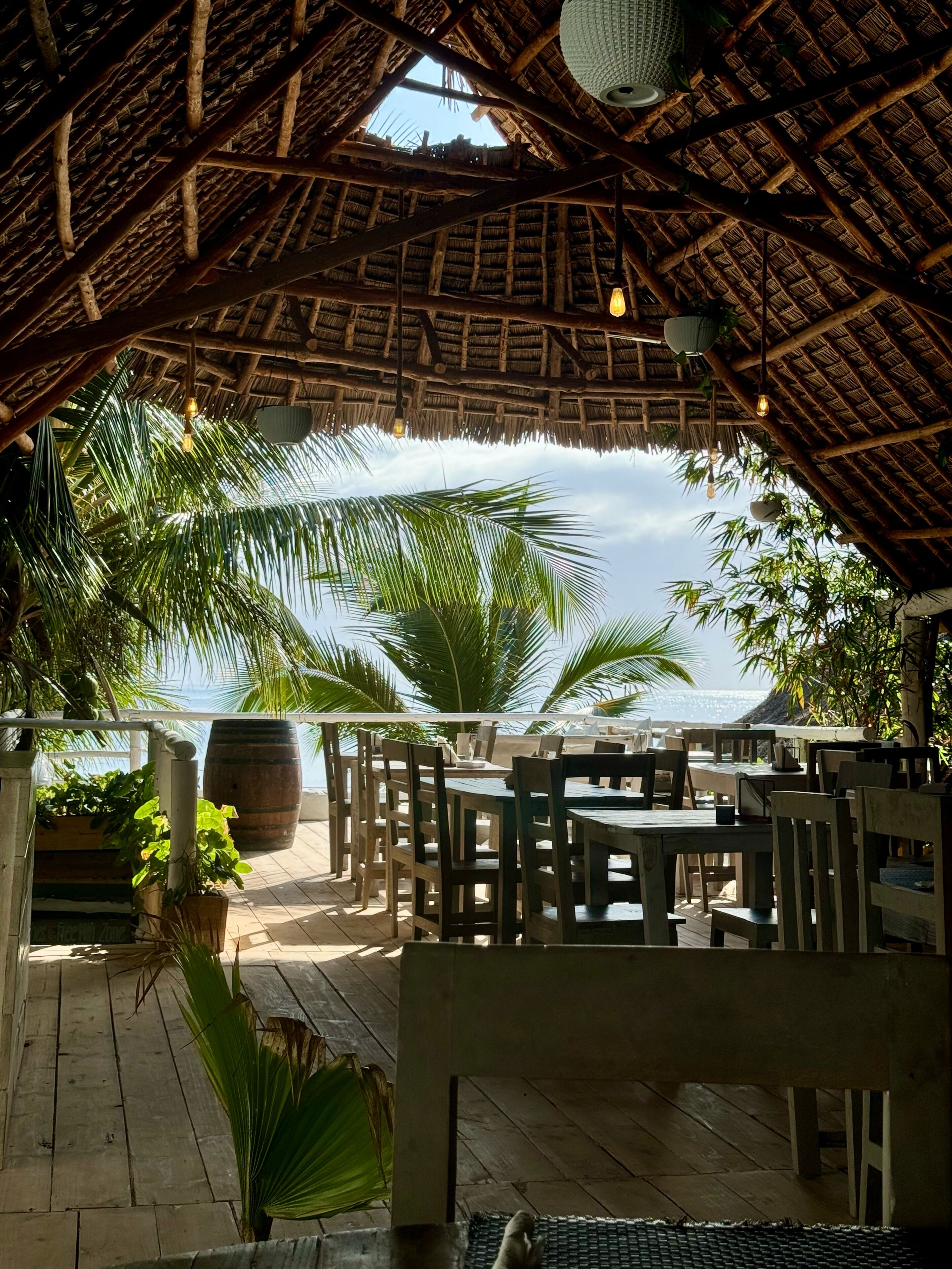 A thatched-roof outdoor dining area with wooden tables and chairs, surrounded by palm trees and a view of the ocean.