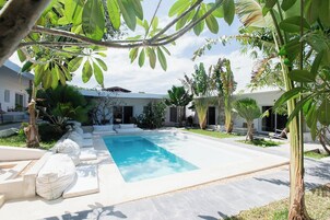 Una piscina rodeada de sillas reclinables blancas, un solárium de hormigón y plantas tropicales.