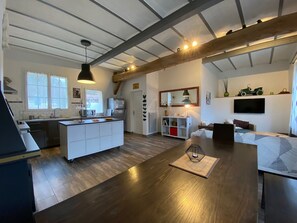 Een moderne keuken met een centraal eiland, een eetgedeelte met een houten tafel, en een woonkamer met een bank en televisie.
