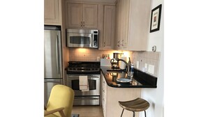 A compact kitchen with stainless steel appliances, a yellow chair, and a bar stool. - Wonderful beachside hideaway. Steps to the beach! Steps to the action! (Provincetown)