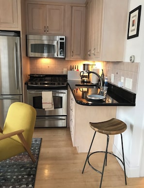 Cocina compacta con electrodomésticos de acero inoxidable, una silla amarilla y un taburete.