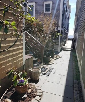 Un étroit sentier extérieur bordé de bâtiments, agrémenté de plantes en pot et d’une clôture en bois.