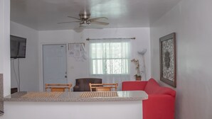 Un espace de vie compact comprenant un coin cuisine, un ventilateur de plafond, un coin repas avec deux chaises, un canapé rouge et une fenêtre avec des rideaux légers.