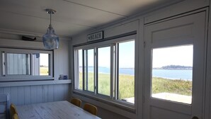 A room with a large window overlooking a body of water, a white door, and a table with chairs. - Popham Beach Direct Oceanfront Cottage Steps to the sand (Phippsburg)