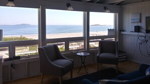 A room with a large window overlooking a beach, two chairs, a small table, and a TV on the wall. - Popham Beach Direct Oceanfront Cottage Steps to the sand (Phippsburg)