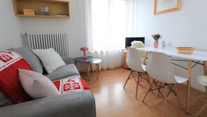 Un salon moderne avec un canapé gris, des coussins blancs et rouges, une petite table ronde, un bureau blanc avec un ordinateur et une fenêtre ornée de rideaux légers.