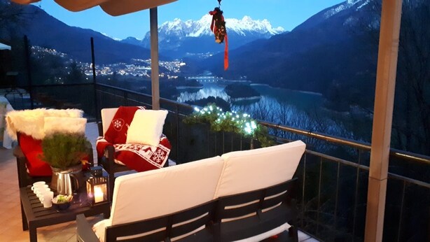 Un balcon offrant une vue sur un lac et des montagnes, aménagé avec un canapé blanc et des coussins rouges et blancs, ainsi qu’une table sur laquelle sont posés une bougie et une plante.