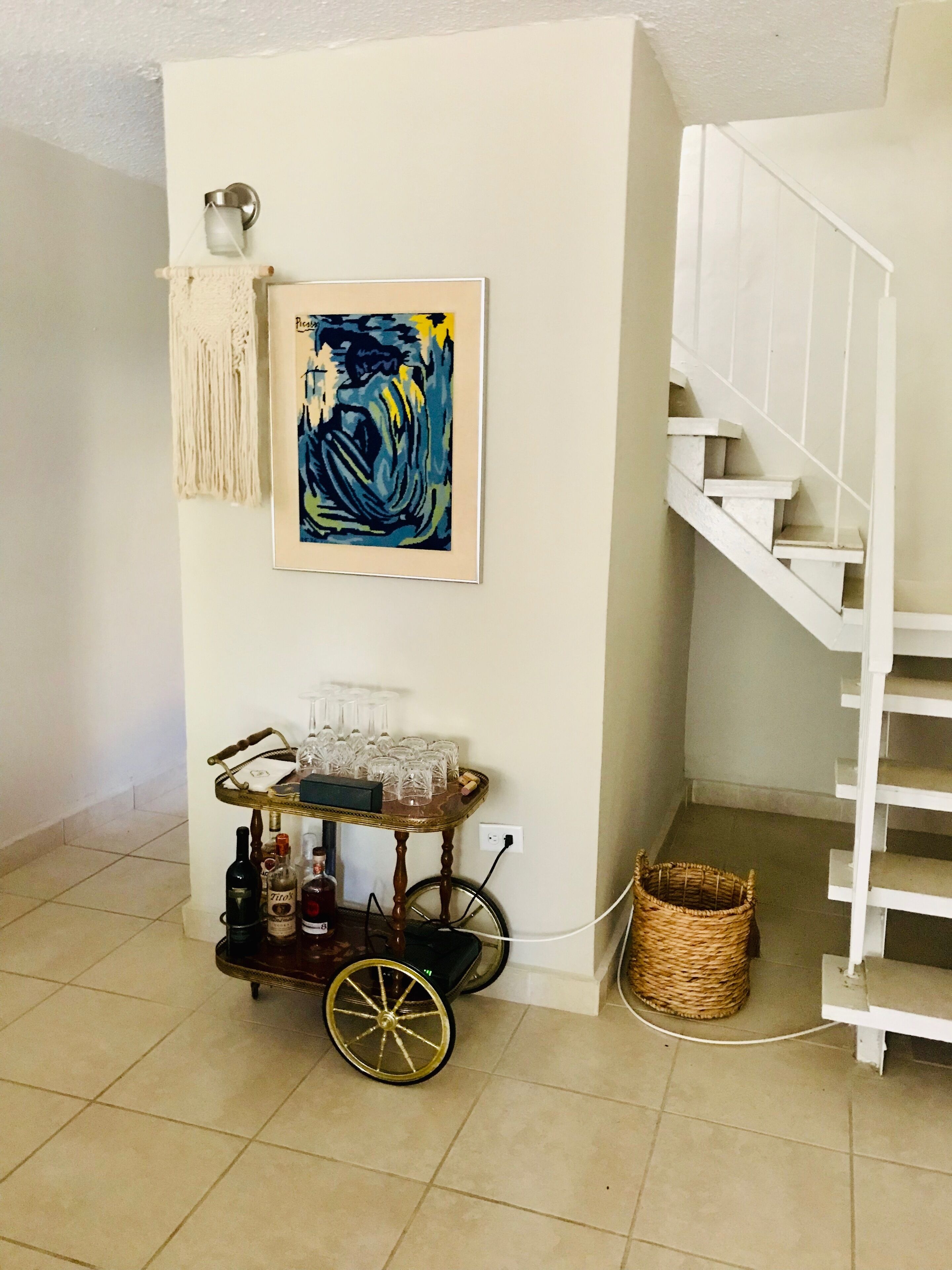 Una habitación con una escalera blanca, un aplique de pared, un cuadro abstracto enmarcado, un carrito pequeño con copas y botellas, y una cesta de mimbre.