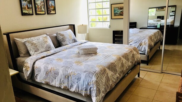 Cama bem arrumada com colcha estampada, mesa de cabeceira com abajur e janela com vista para o verde.