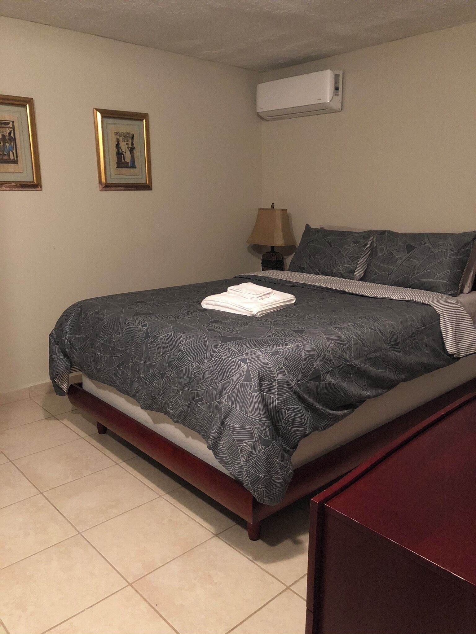 Una cama bien hecha con una colcha estampada oscura y almohadas, una lámpara de noche y dos cuadros en la pared.