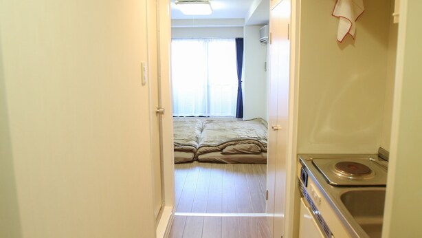 A narrow hallway leading to a bedroom with a bed and a window. A kitchen area with a sink and stove is visible to the right. - Kanazawa-Hub (Kanazawa)