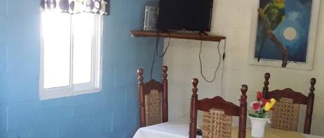 A dining area with a table and chairs, a television on a shelf, a window with a curtain, and a painting on the wall.