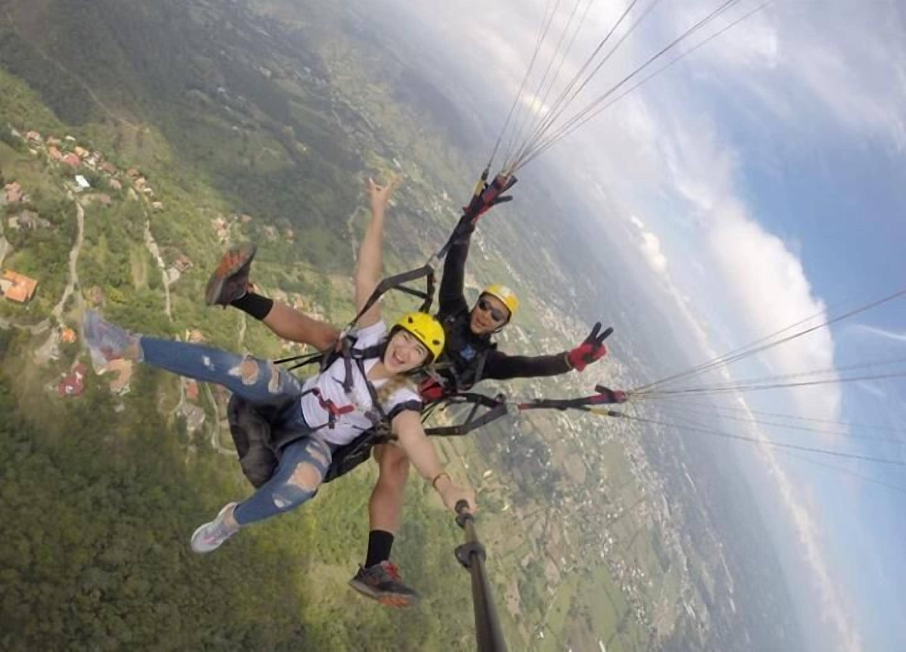 Two individuals skydiving with parachutes, surrounded by a mountainous landscape and scattered houses.