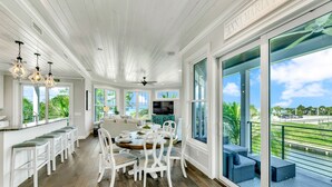 A spacious dining area with a large round table and white chairs, a kitchen area with a TV, and a balcony with a view of the water.